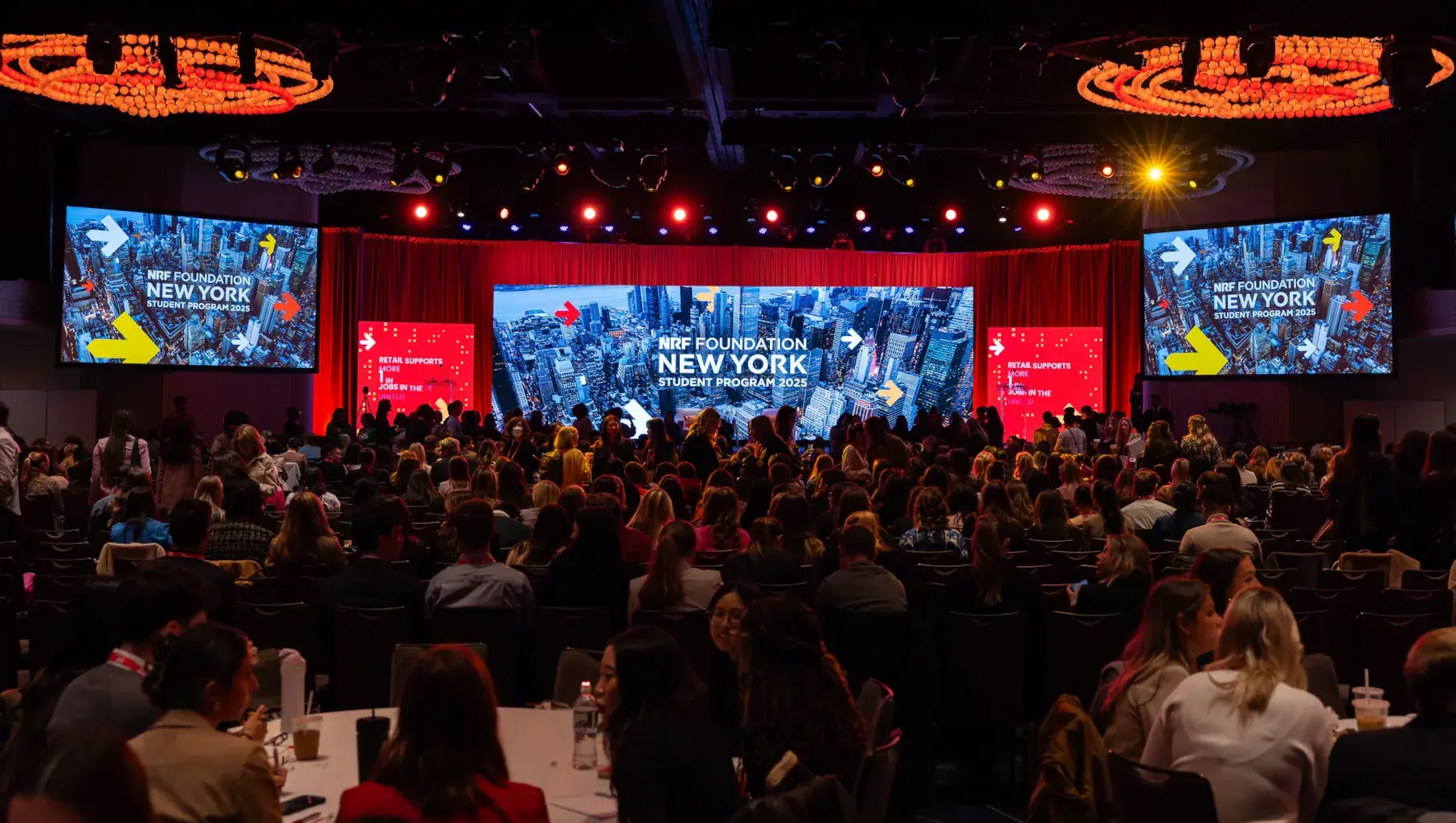 Hundreds of students and graduates are seated in eager anticipation of the start of the 2025 NRF Foundation Student Program. In the foreground, students discuss retail topics with one another while in the background, giant screens perched on a stage cycle through branded images of New York and elements of the Student Program.